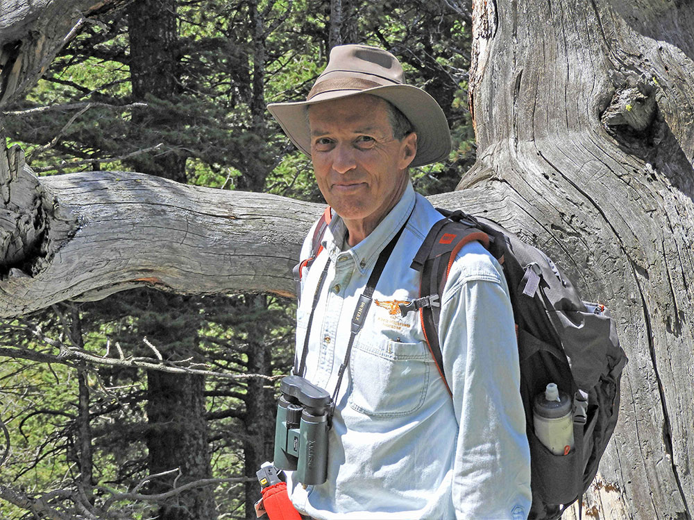 A man in a white shirt and hat stands by the branch of a tree with a pair of binocoulars hanging around his neck.