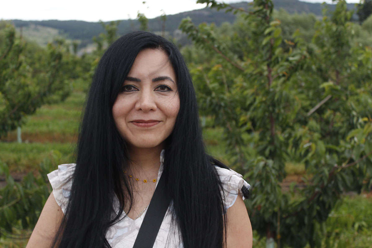A woman with long black hair and a white blouse smiles at the camera while standing in an orchard.