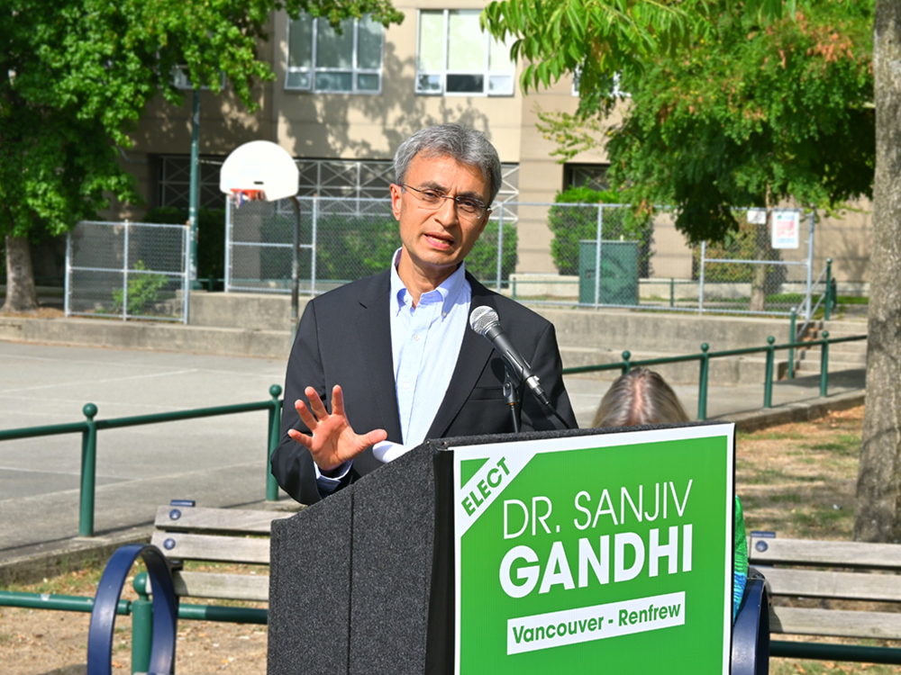 Sanjiv Gandhi, wearing a blue collared shirt and suit jacket, speaks behind a podium with a sign that reads ‘Elect Dr. Sanji Gandhi, Vancouver-Renfrew.’