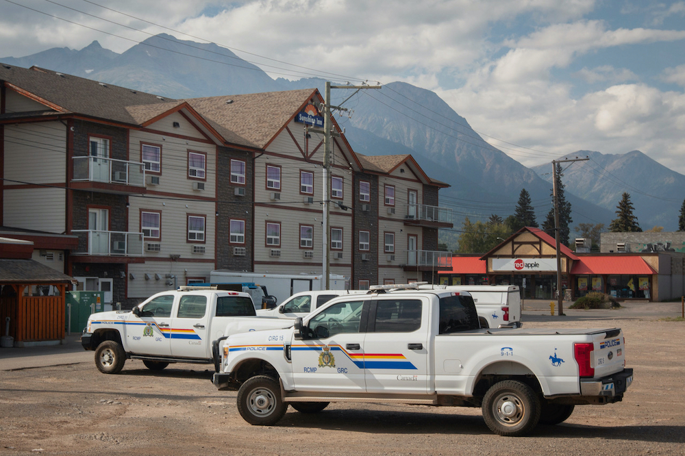Three white pickup trucks with police markings on the side are parked in front of a three-storey building with mountains behind.