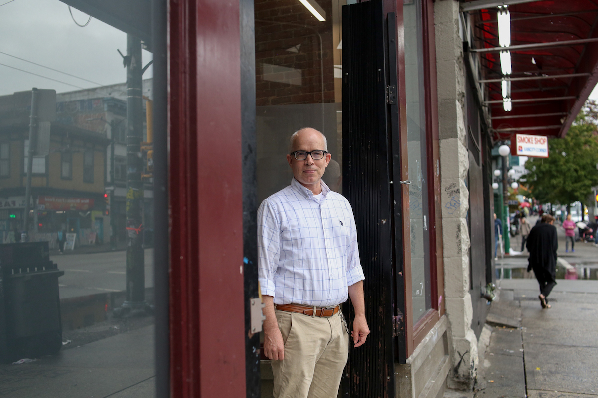 A man with rolled-up sleeves and glasses stands outside a doorway in a city.