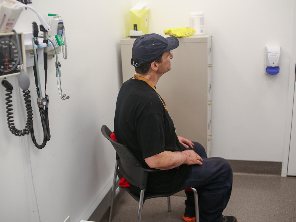 A man sits on a chair, semi-turned away from the camera, in a doctor’s office.