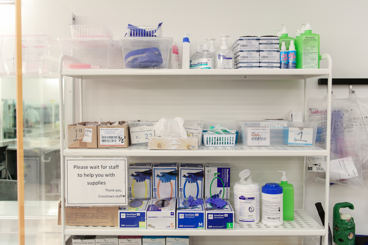 Harm reduction supplies, such as latex gloves, are arranged on a shelving unit. There is a sign saying, “Please wait for staff to help you with supplies.”