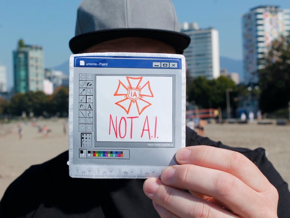 Jo Stilwell stands on a Vancouver beach and holds a sign that says “IA not AI.” His face is hidden.