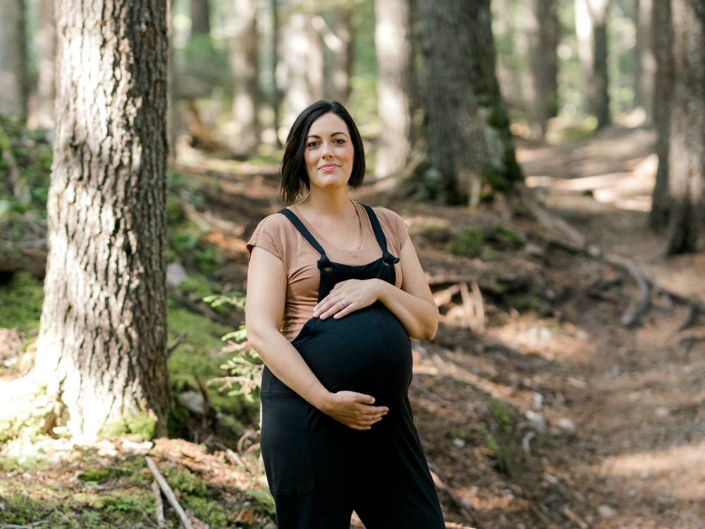 A woman wearing black overalls stands in a forest, her hands on her belly.