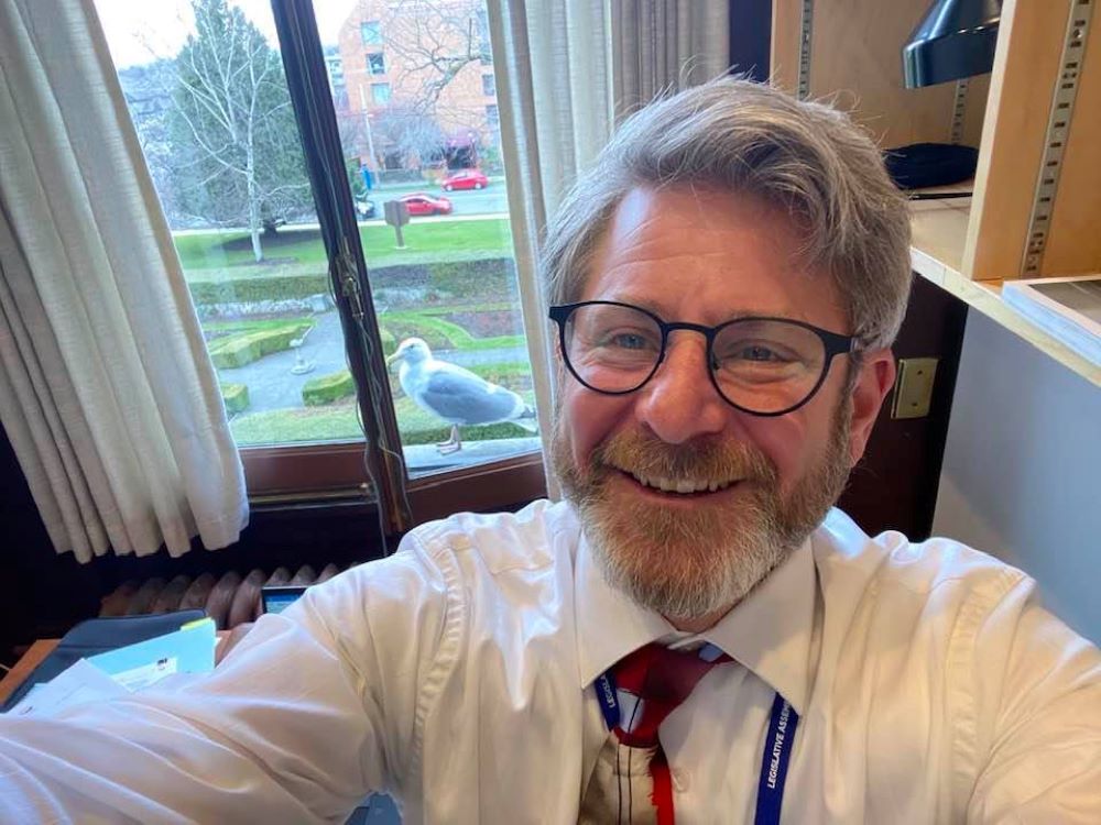 Bruce Banman in white shirt and tie takes a selfie in his legislature office, with a seagull looking in from the windowsill.