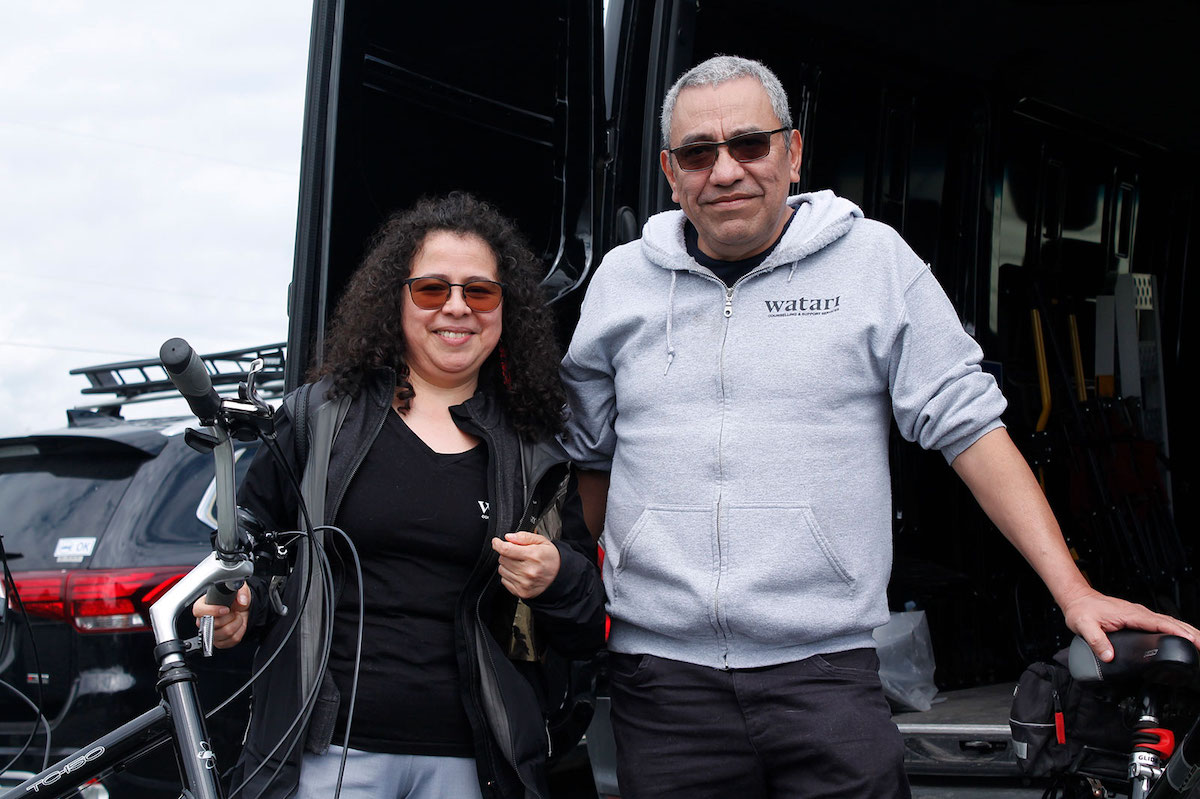 Byron Cruz and Ingrid Mendez de Cruz stand in front of an open Sprinter van, their hands resting on two bicycles.