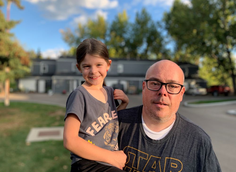 A bald man with glasses and wearing a dark grey shirt that reads "Star Wars" carries a smiling girl with a t-shirt that has a tiger on it and reads "Fearless." Behind them are trees and rows of grey houses.