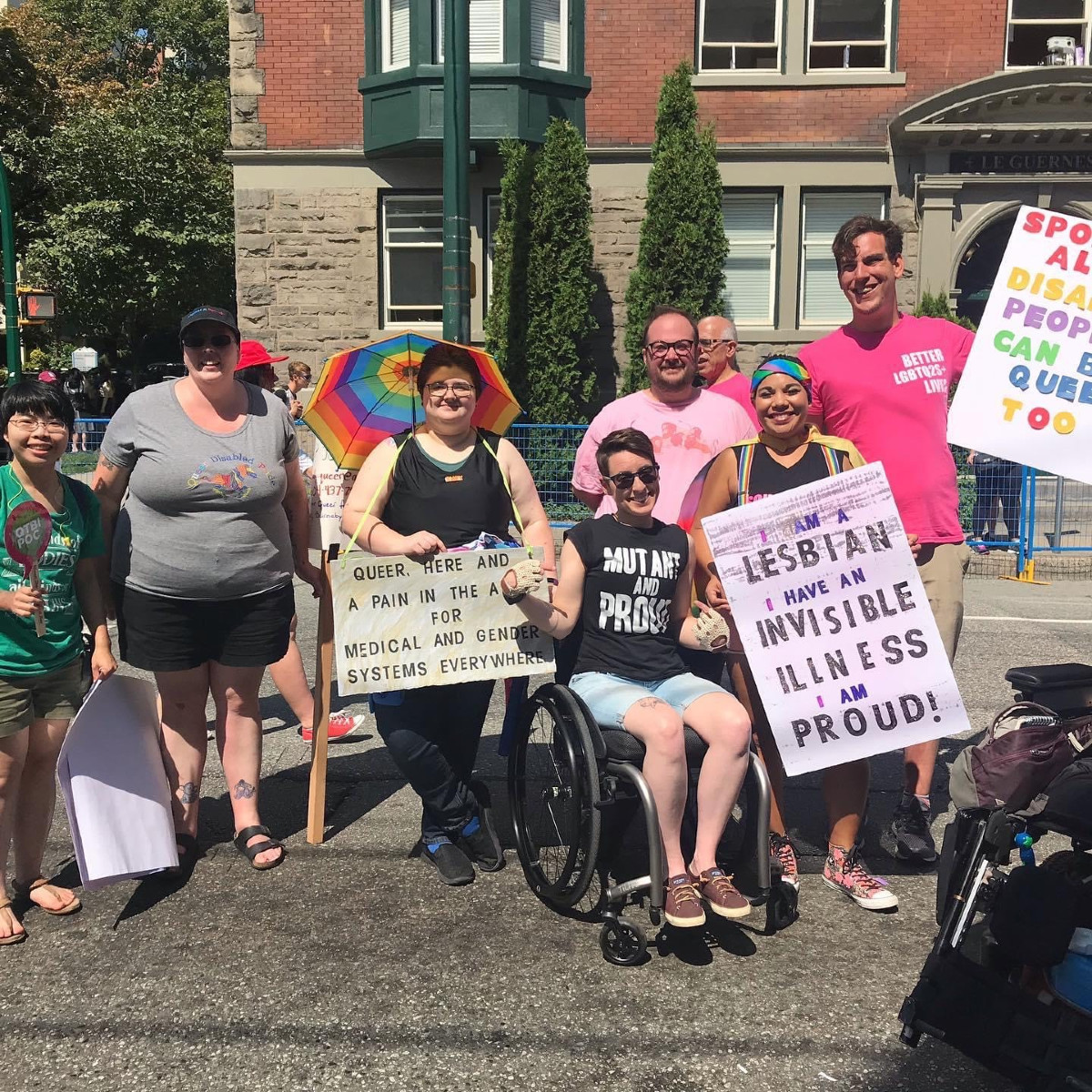A group of people gather near Davie Street at Vancouver Pride, holding placards. One says, ‘Queer, Here, and a Pain in the Ass for Gender and Medical Systems Everywhere,’ and another says, ‘I Am a Lesbian. I Have an Invisible Illness. And I am Proud.’