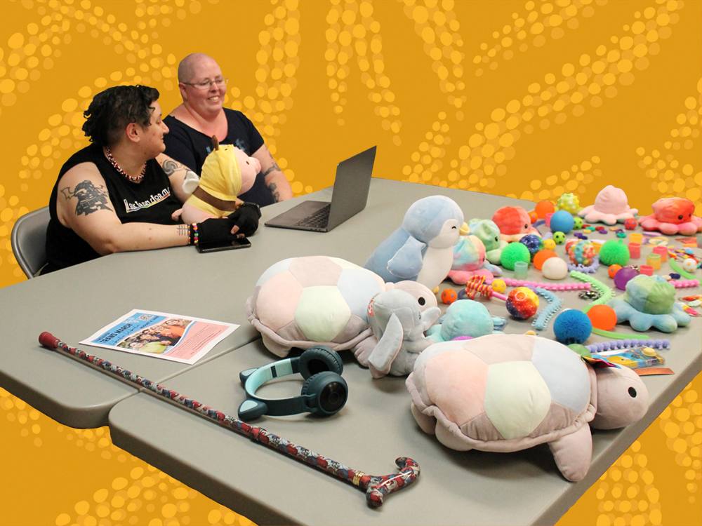 Two people sit at a table that features a cane, noise-cancelling headphones, and a variety of plush animals and stim toys.