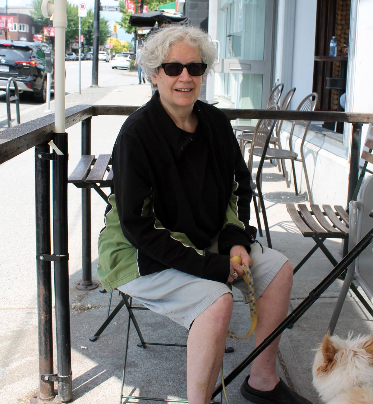 A woman sits on a patio. A small dog on a leash sits on the pavement, looking at her.