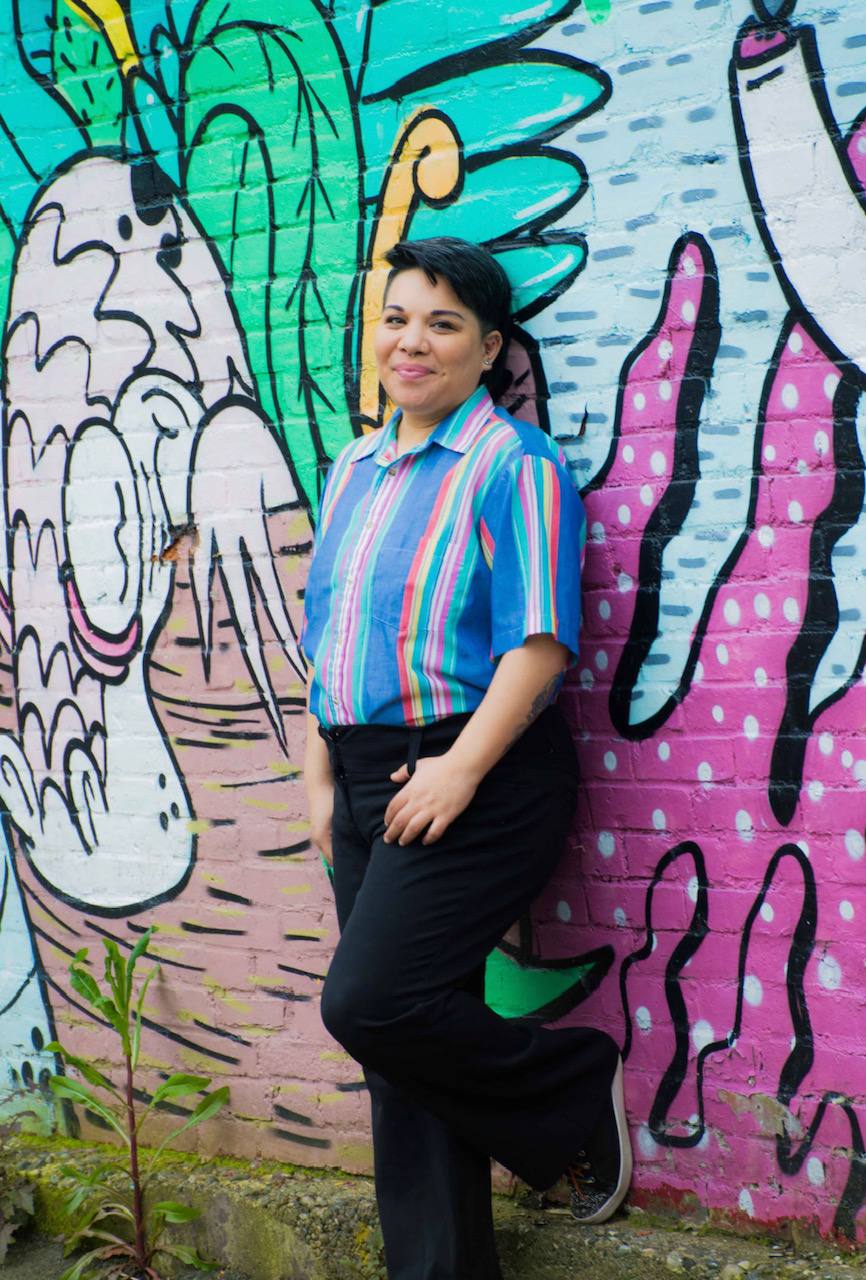 A woman in a colourful striped shirt and black jeans leans against a wall with a mural.