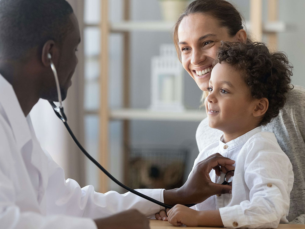 A doctor holds a stethoscope against a child’s chest. The child is sitting in a woman’s lap.
