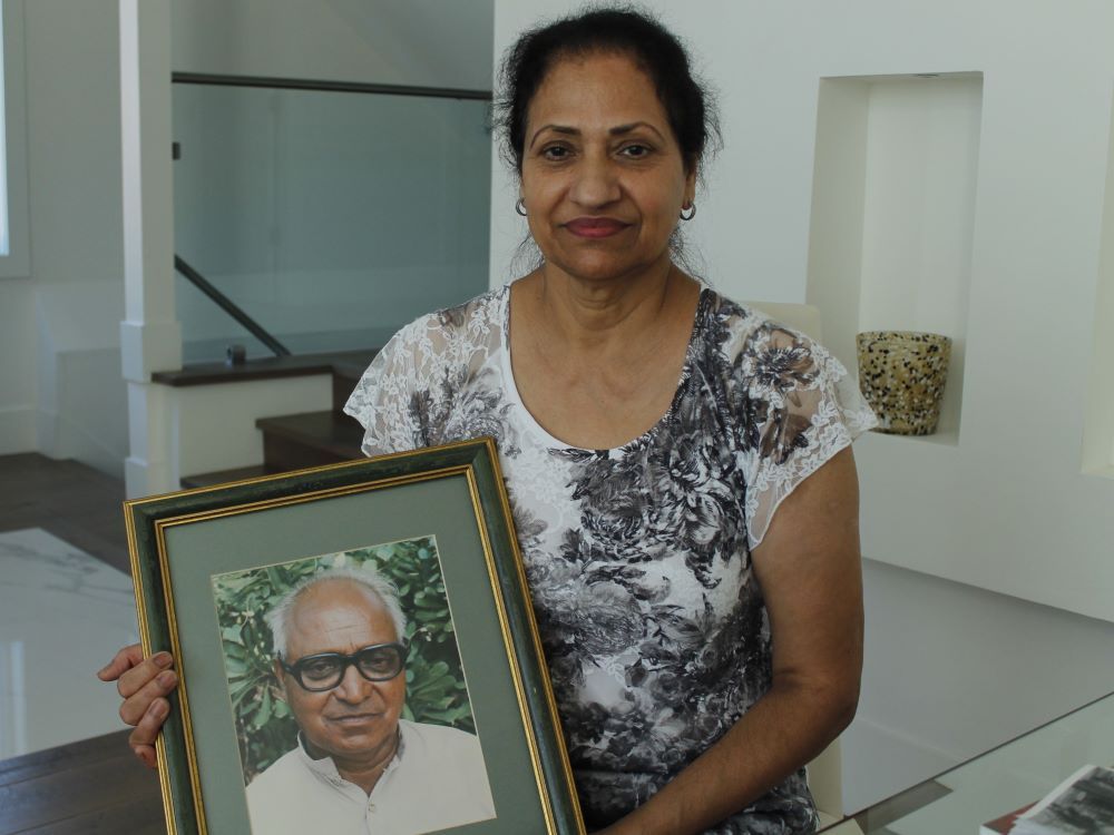 A South Asian woman stands indoors holding a portrait of her father, a balding older man with white hair and large, black-framed glasses.
