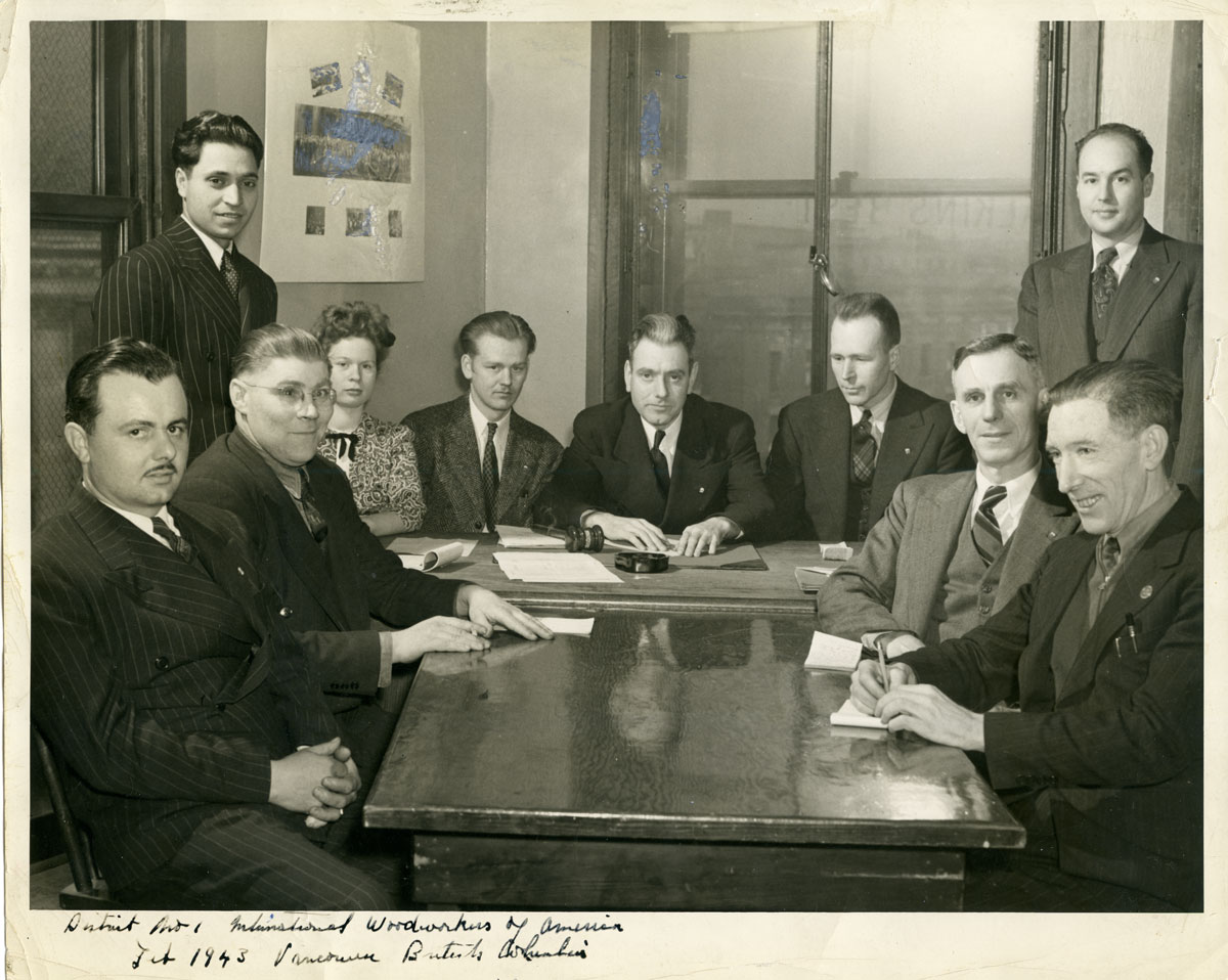 A group of men and a woman congregate around conjoined solid wood tables and stare into the camera, some smiling and others straightfaced. To the left of the image, wearing a pinstriped suit with a tie is Darshan Singh Canadian.