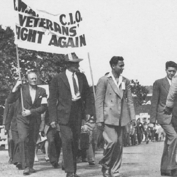 Suited men march in a column, some holding up a banner that says ‘IWA CIO Veterans Fight Again.’ At the centre of the image is a South Asian man, Darshan Singh Canadian, wearing a light-coloured suit without a tie.
