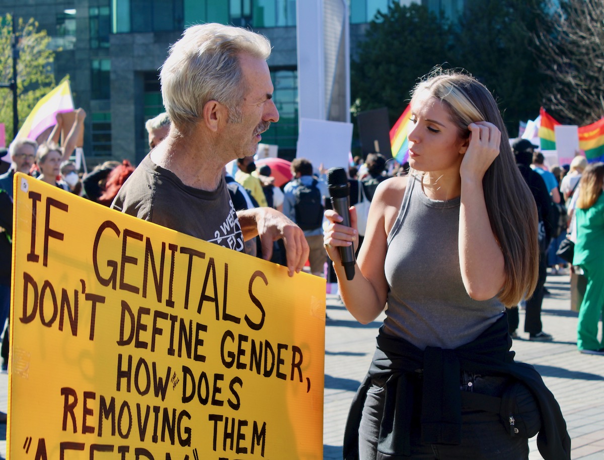 On the left, a man holds a placard with an anti-trans message. He speaks into a microphone held by Lauren Southern, on the right.