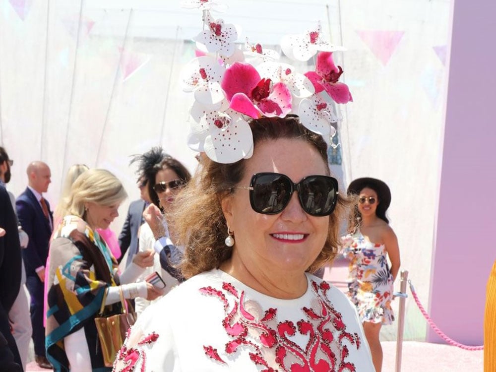 A smiling woman, global mining mogul Gina Rinehart, wears sunglasses and a headdress made of flowers at a horse racing event in Australia.