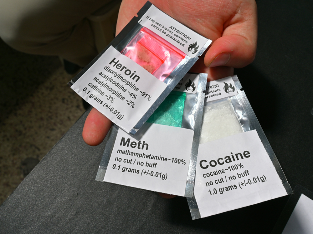 A person holds three baggies. One contains heroin, one contains meth and one contains cocaine. The breakdown of substances in each baggie is listed in a label on the bag. For example: Cocaine: 100%. No cut/no buff.