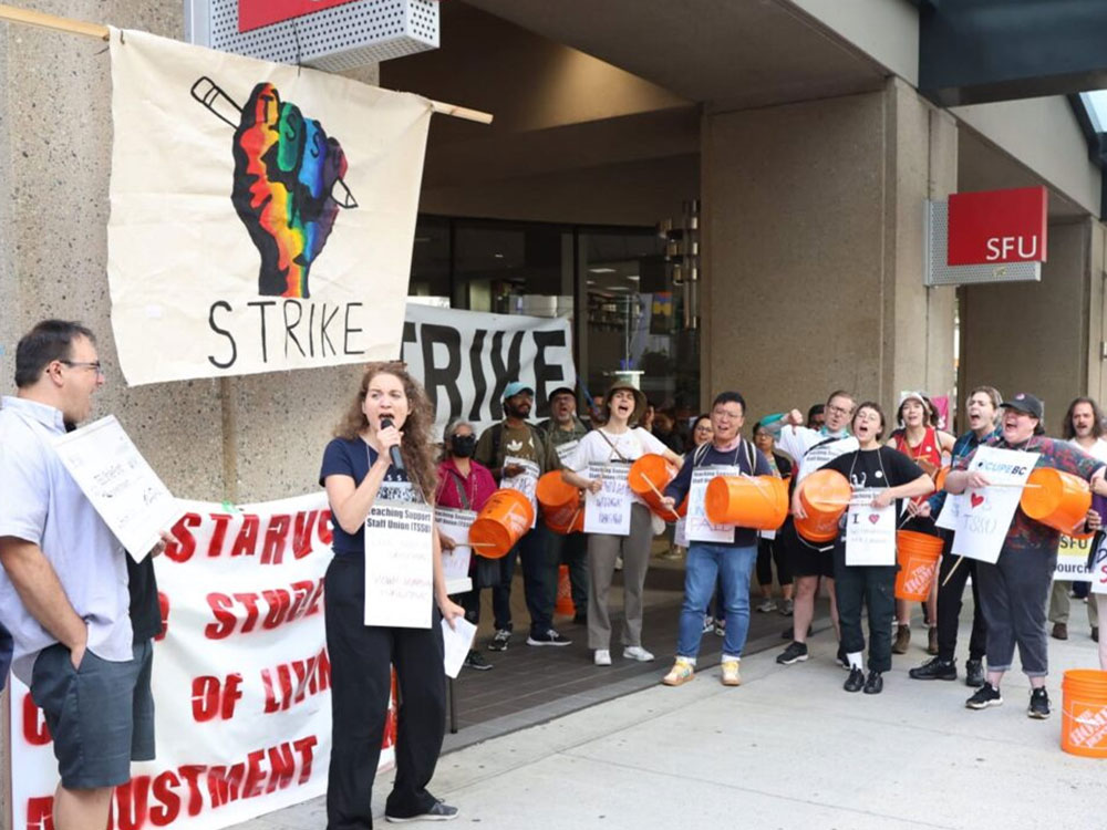 A person holds a microphone under a sign that says "Strike." Others hold placards and drums.
