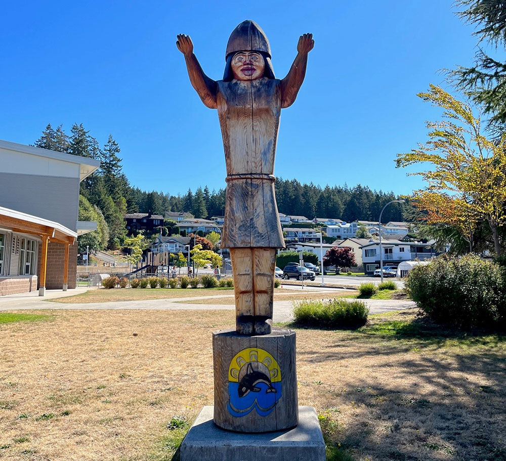 A carved wooden pole features a figure with their arms up in a welcome gesture. In the background, a playground and the entrance to an elementary school are visible.