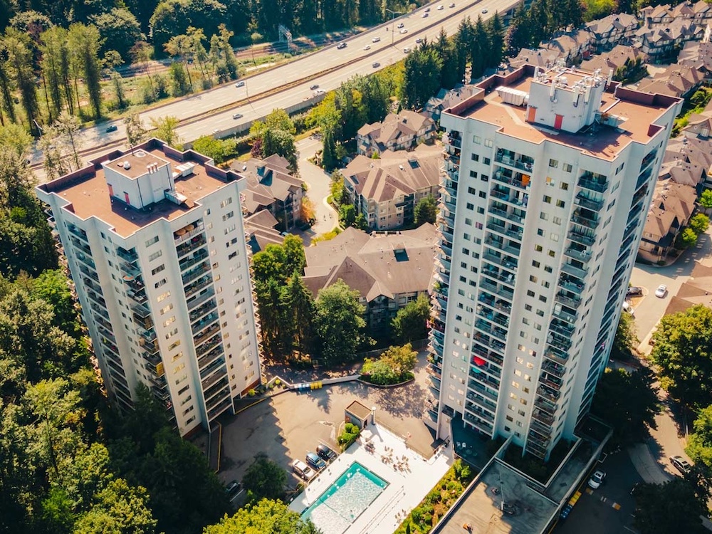 An aerial photograph features two highrise residential towers surrounded by lush green stands of trees and blocks of low-rise townhomes with brown roofs. It’s a sunny day.