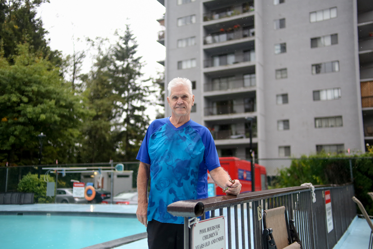 A man with white hair, a blue exercise shirt, sporty shorts and flip-flops holds a pair of keys while he leans against a fence. Behind his right is an outdoor pool. Behind his left is a concrete apartment tower.