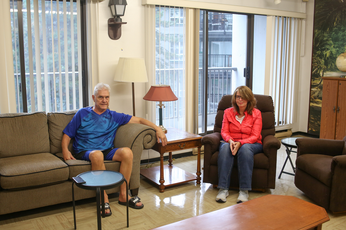 A senior man and a woman sit on old couches of a 1980s apartment tower social room. It looks cosy.