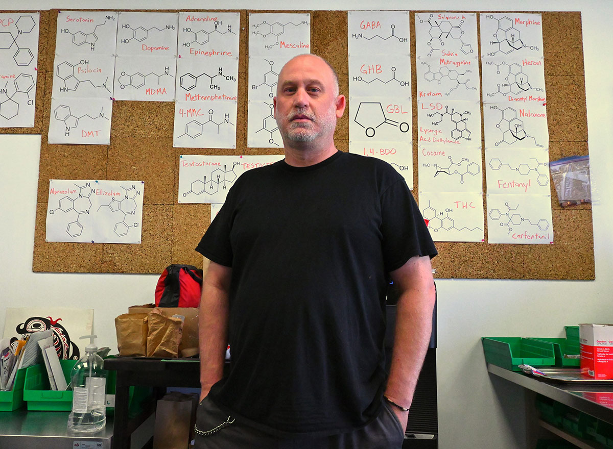 A man in a black T-shirt stands with his hands in his pockets. On the cork board wall behind him are pieces of paper with the molecules of different substances drawn on them.