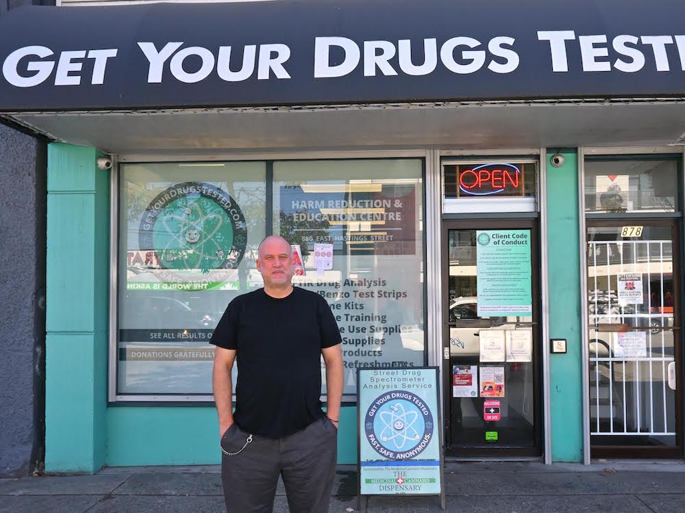 A white man in a black T-shirt stands out front of a teal-coloured store. The black awning reads “Get Your Drugs Tested.”