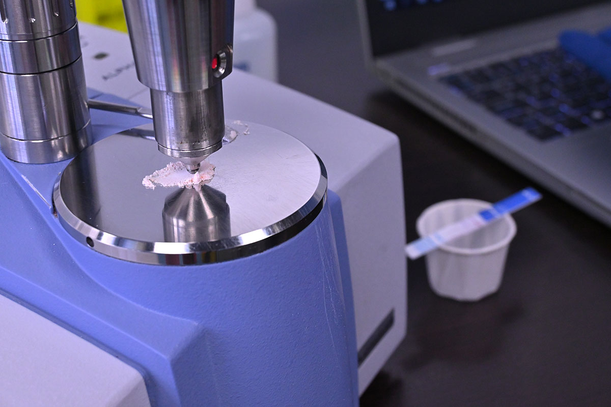 A small sample of white powder sits on a silver plate under a sensor on a blue and grey machine. In the background two white and blue test strips rest on a paper portion cup.