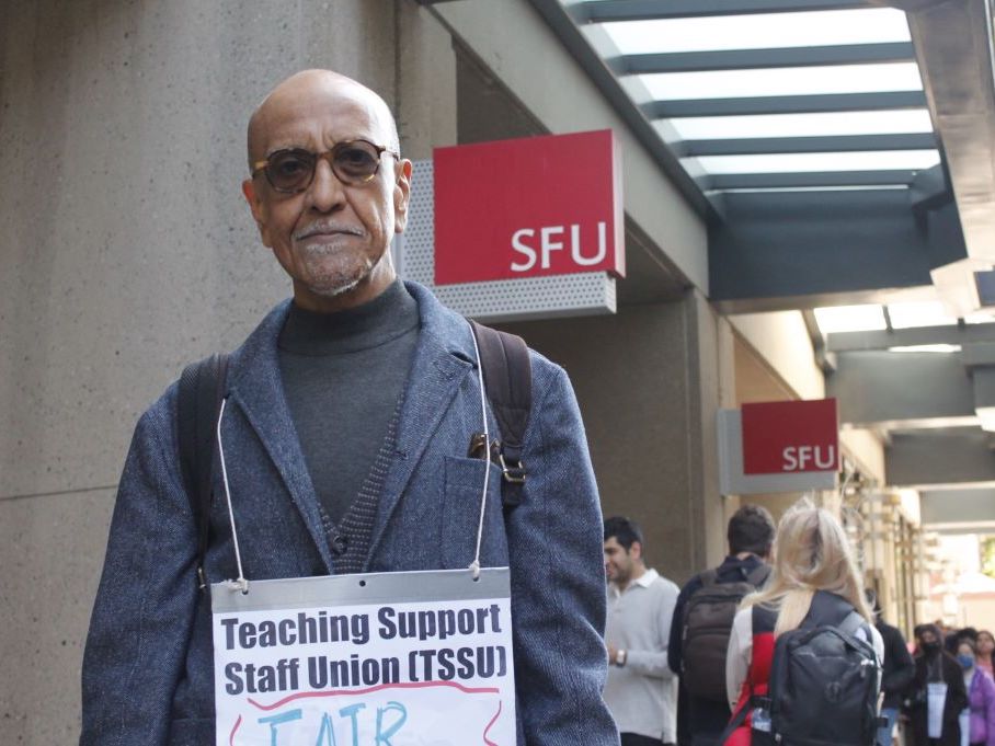 A man in sunglasses wearing a sign that reads “Teaching Support Staff Union (TSSU)” stands in a picket line outside an SFU campus.