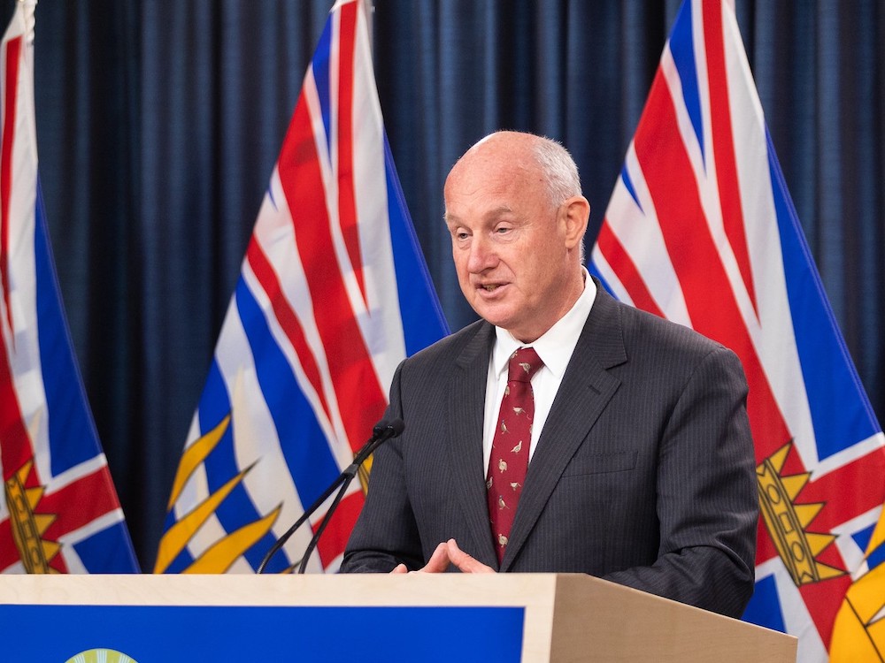 An older man stands at a podium and appears to speak. Behind him are flags for the province of B.C.