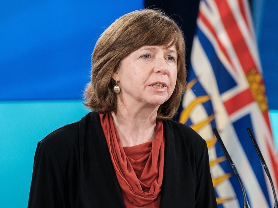 A woman with brown hair wearing a black cardigan over a red blouse stands at a podium speaking. A British Columbia flag is visible behind her.