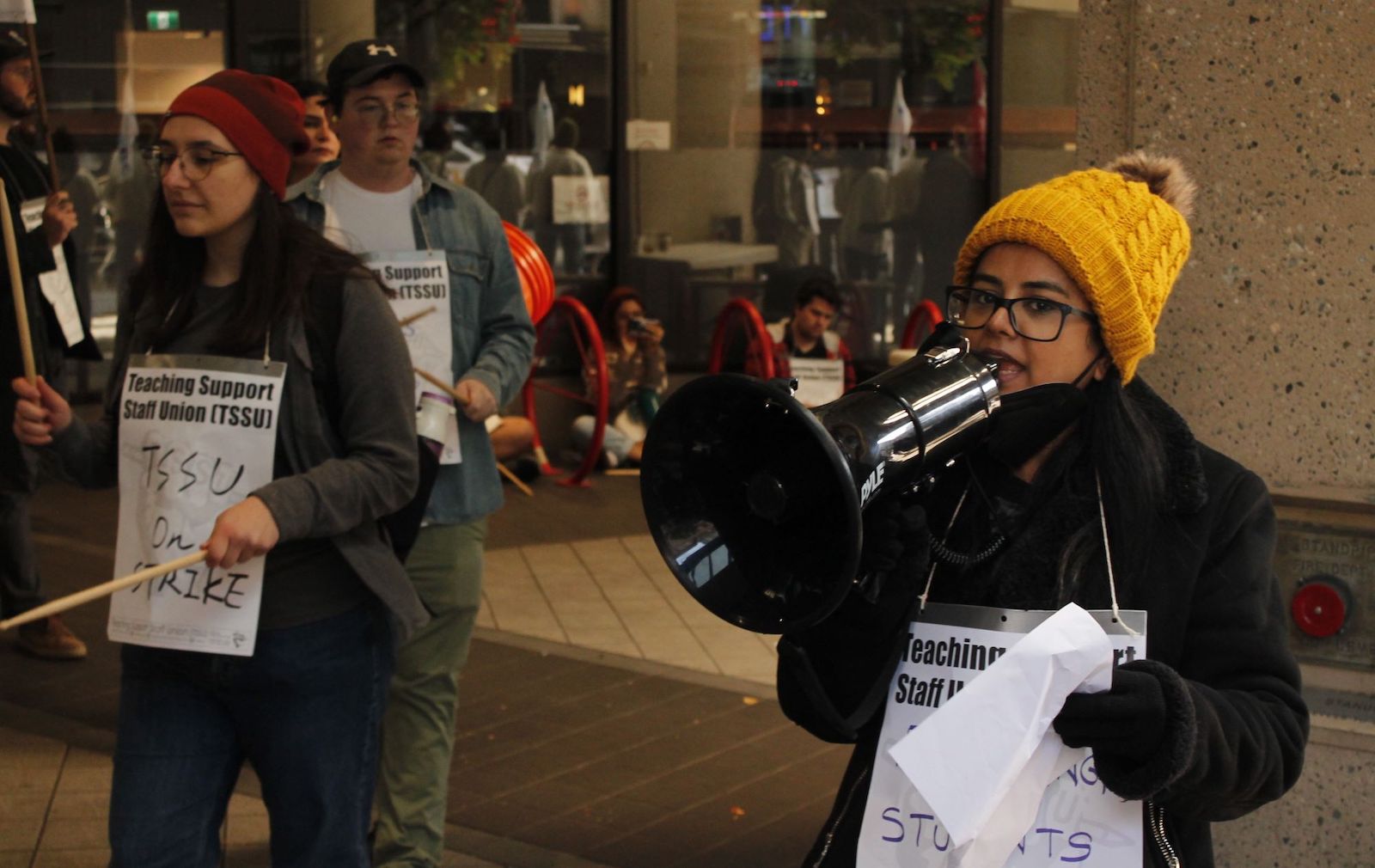 In the foreground, a woman in a yellow toque and glasses speaks into a megaphone. To her right, another woman holding a drumstick in each hand wears a red toque, glasses and a sign that reads “Teaching Support Staff Union: TSSU on strike.” More picketers can be seen in the background.