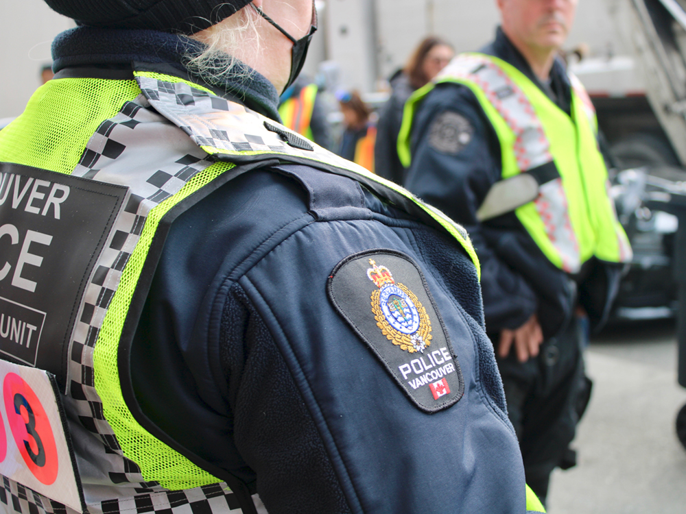 Two police officers are visible. One stands with their back to the camera, wearing a medical mask. The other is facing partway towards the camera. Both are wearing yellow safety vests.