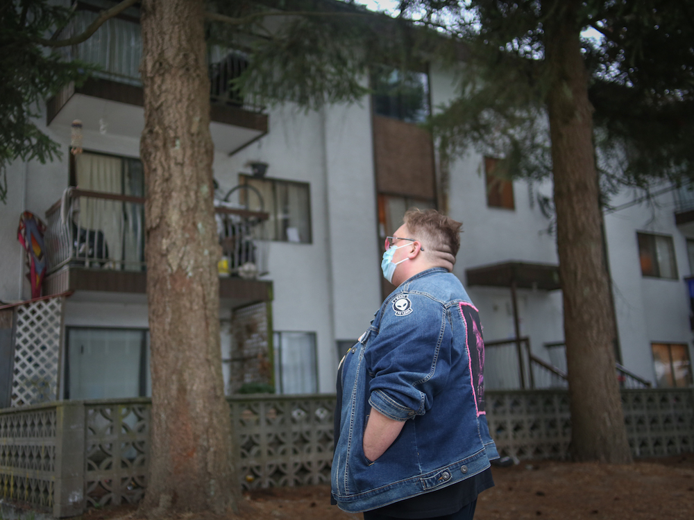 A person stands in front of a wood-frame rental walk-up building. You can see the balconies and there is a lot of stuff on them, giving it a lived-in feel.