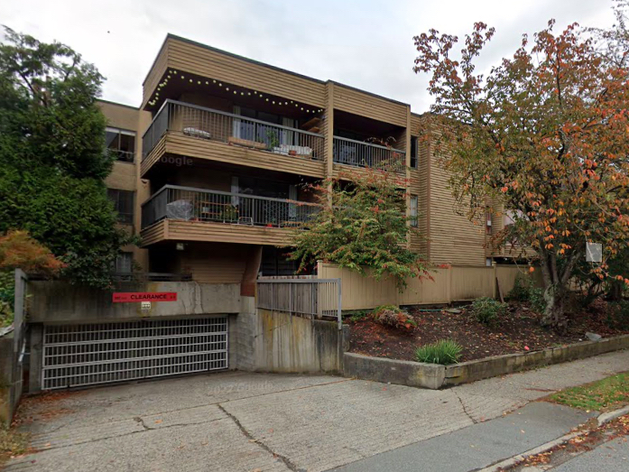 A three-storey walk-up in Vancouver appears to be from the mid-’70s. The siding is brown, the balconies are metal. Cracked concrete leads to an underground garage in the foreground.