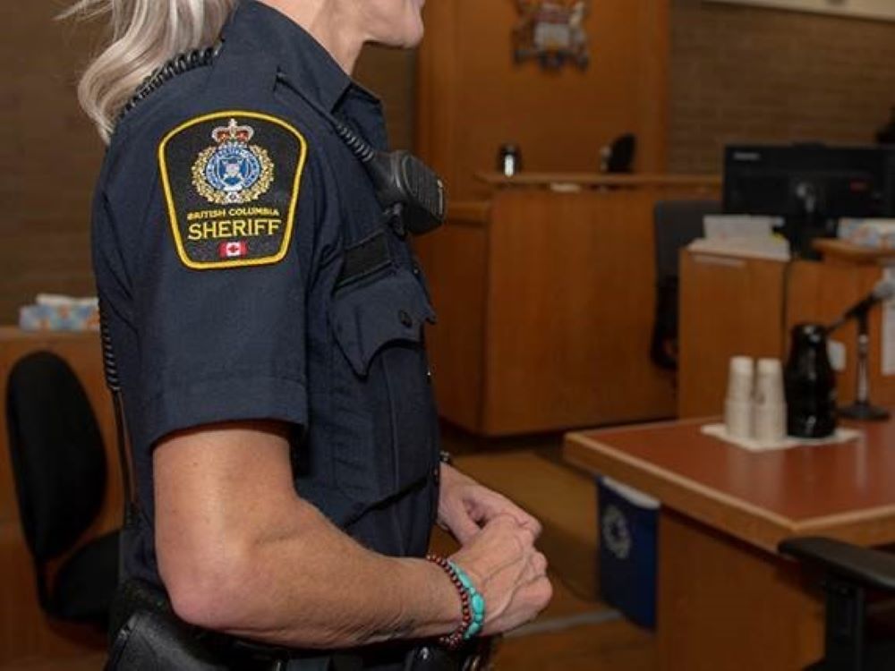 A blond sheriff in a navy-blue uniform stands in a courtroom.