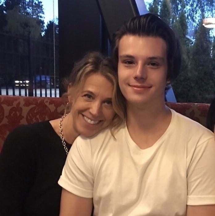 A woman with blond hair and a black top leans to the right, into the shoulder of her young adult son. He is wearing a white T-shirt and has dark hair. They are sitting in a red upholstered restaurant booth and the window behind them overlooks a quiet street.