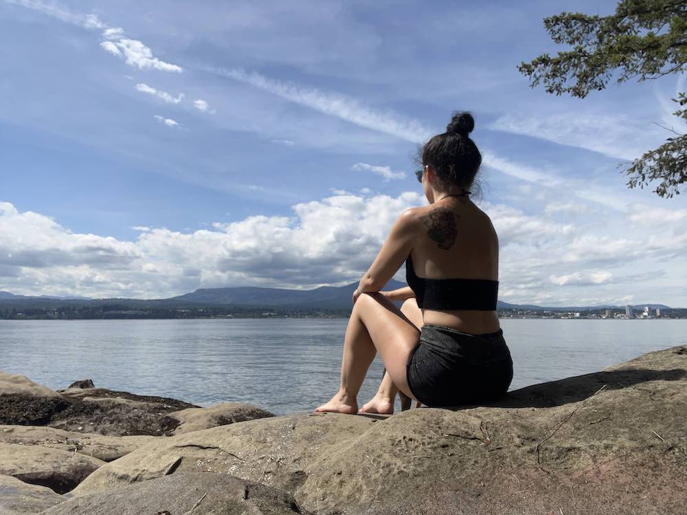 A woman sits on a rock with her back turned to the camera, looking out to the water in front of her. She has her black hair tied up in a high bun. She is wearing black shorts and a black crop top. A tattoo is visible on the back of her left shoulder.