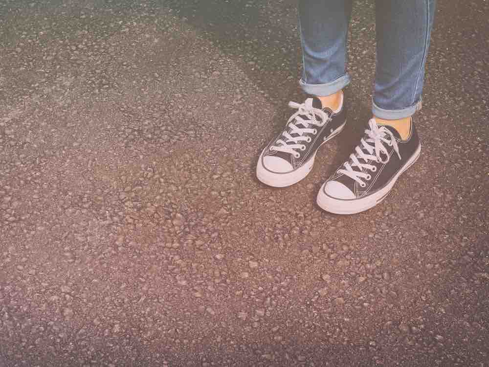 Shot from the calves down, a person wears youthful, straight-legged blue jeans with their cuffs rolled up over low-rise black Chuck Taylor sneakers with white laces. They are standing on black asphalt.