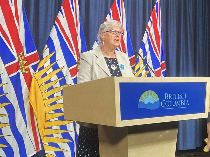 A blonde white woman wearing glasses, a polka dot blue shirt and light-coloured blazer stands at a podium in front of B.C. flags. The podium says 'British Columbia' with the province's logo.