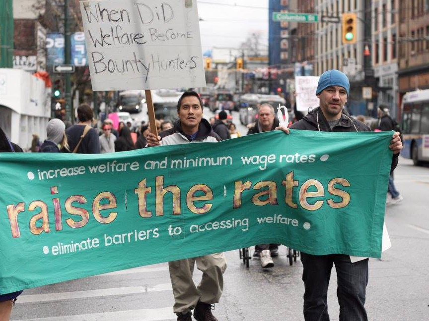 Two people carry a green banner that says “Raise the Rates” on a Vancouver street under grey skies.