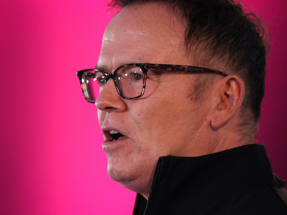 A closeup photo of a white man with short brown hair, wearing glasses and a high-collared black jacket. The background is soft purples and reds.