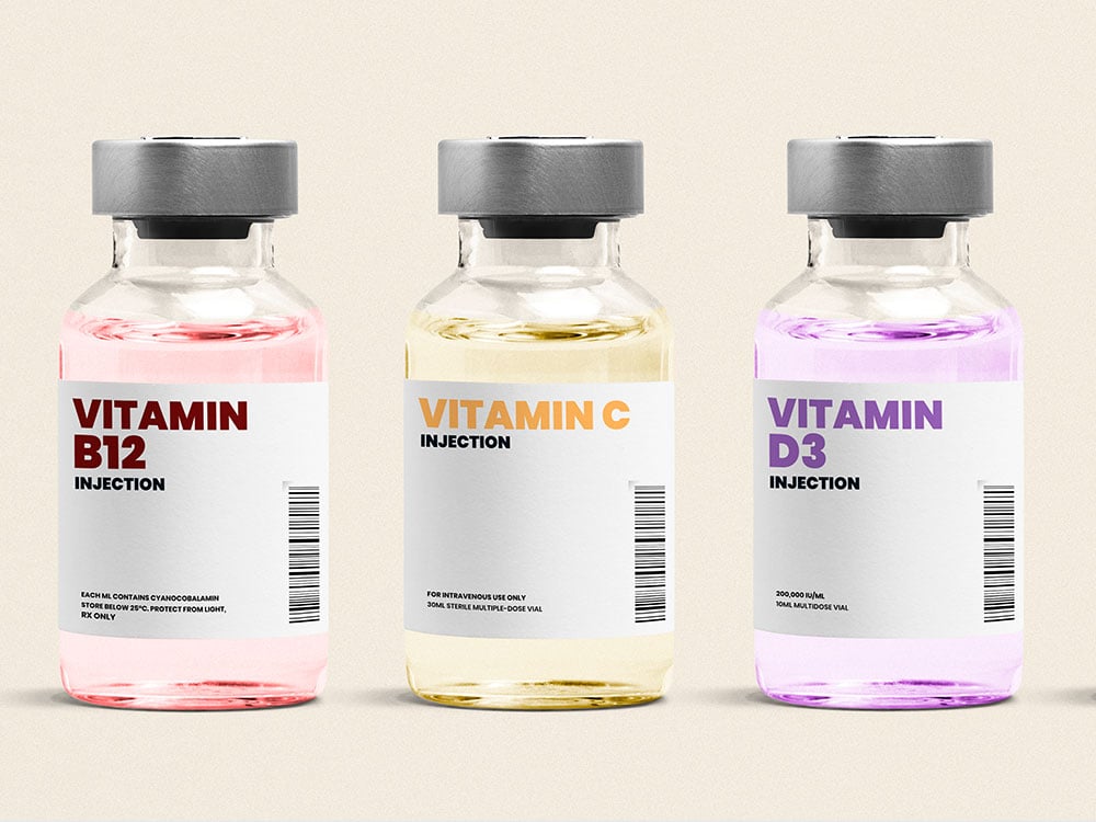 Three vials of vitamin solutions intended for intravenous injection are in a row in front of a beige background. They read "Vitamin B12," "Vitamin C" and "Vitamin D3."
