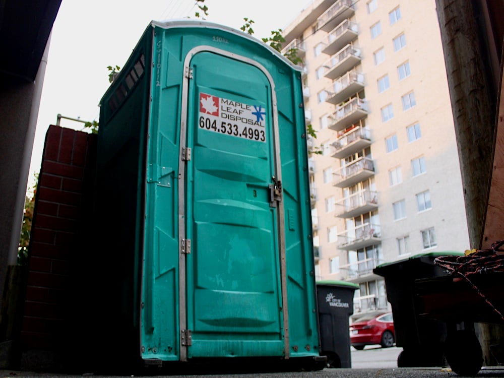 A green porta-potty is in an alley, next to a brick wall. Behind it, there are garbage cans, and, in the background, a West End apartment building.