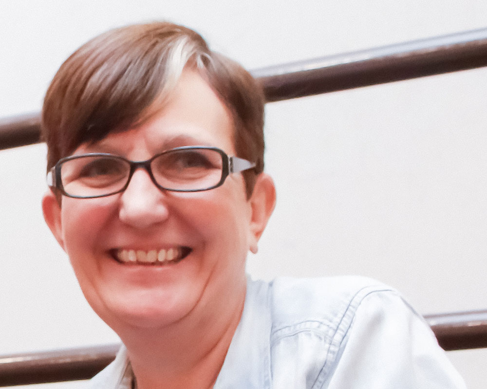 A woman with short hair and glasses smiles at someone behind the camera.