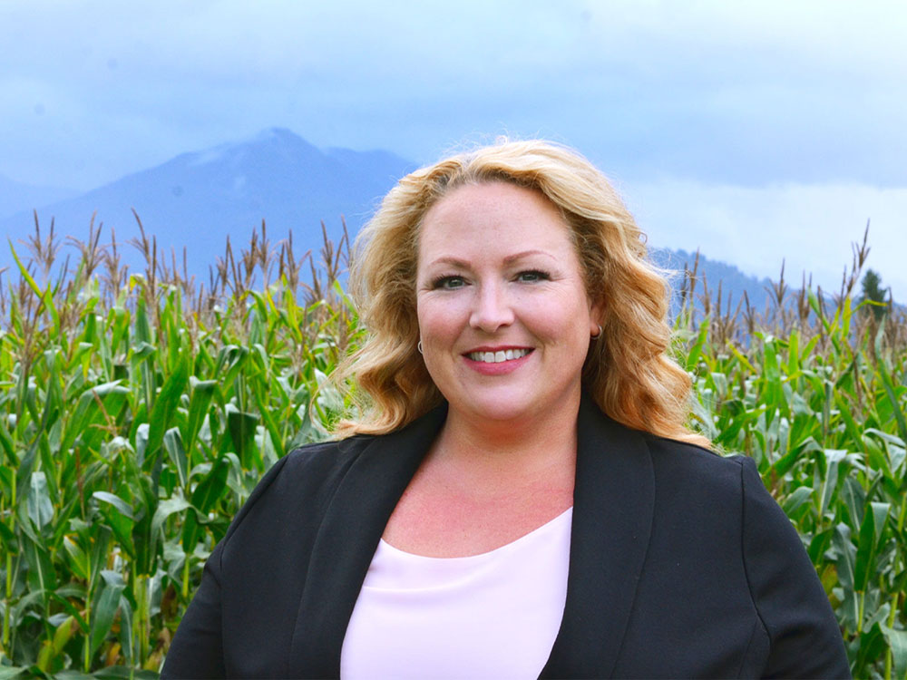 A woman with blond hair, a blue jacket and white shirt stands in front of a cornfield.