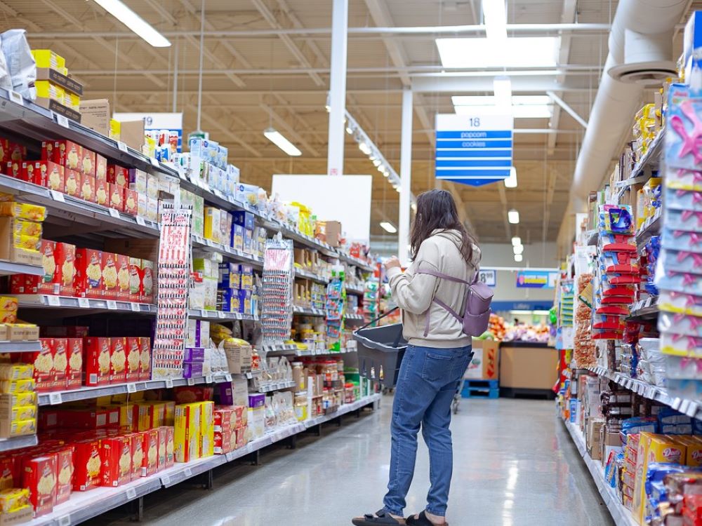 A youth stands in the aisle of a grocery store with a fairly empty basket, unsure of what to choose.